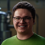 Ein lächelnder Mensch mit Brille und grünem T-Shirt. Im Hintergrund sind unscharfe technische Geräte zu erkennen.