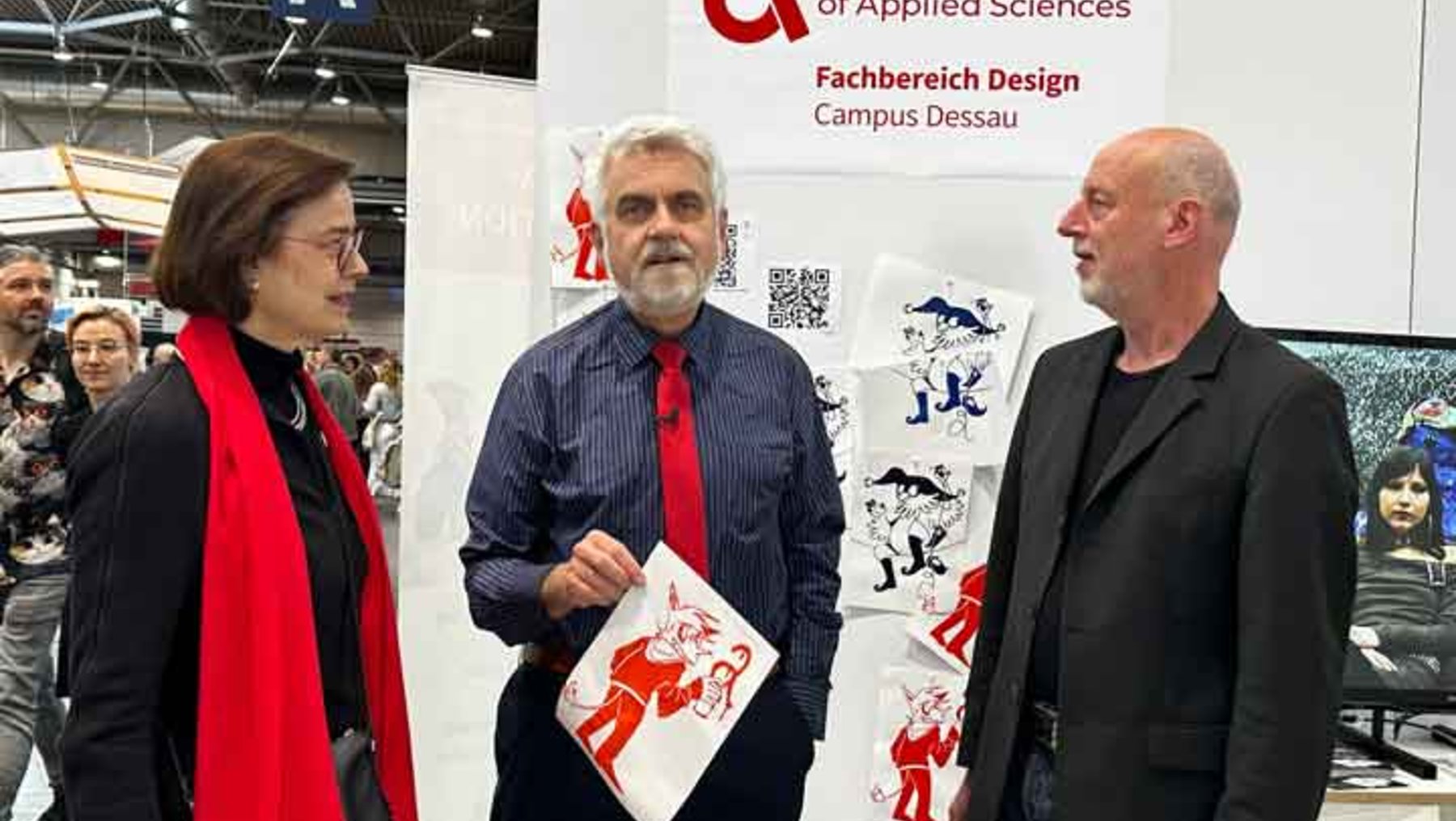 Drei Personen auf einer Messe: Eine Frau in rotem Mantel, ein Mann in blauem Hemd mit roter Krawatte, der ein Poster mit einer schwarzen Zeichnung hält, und ein Mann in schwarzem Anzug stehen vor einem Stand der Hochschule Anhalt, Fachbereich Design, Campus Dessau.