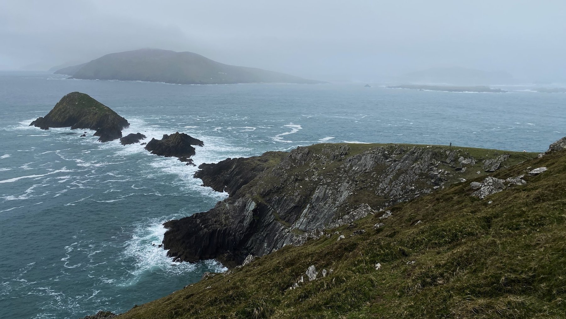 Blick auf eine wilde, nebelverhangene Küstenlandschaft mit steilen Felsformationen, die ins Meer ragen. Im Hintergrund eine größere, ebenfalls nebelumhüllte Insel. Brandung schlägt gegen die Klippen, grüne Grasflächen auf den Hügeln.
