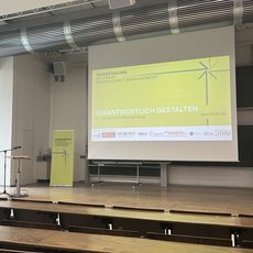 Ein Mann steht auf einer Bühne mit Mikrofon und hält einen Vortrag. Hinter ihm ist eine große Leinwand mit dem Titel „Verantwortlich gestalten – Nachhaltiges Facilitymanagement“ und einem Windrad-Symbol zu sehen. Die Veranstaltung findet in einem hellen Saal mit leeren Sitzreihen statt.