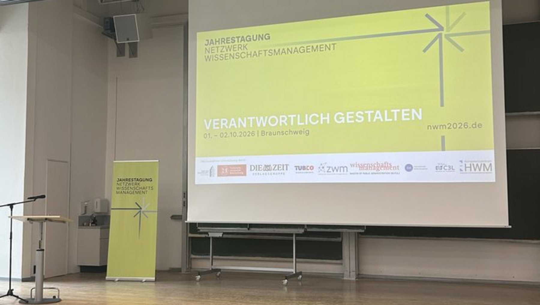 Ein Mann steht auf einer Bühne mit Mikrofon und hält einen Vortrag. Hinter ihm ist eine große Leinwand mit dem Titel „Verantwortlich gestalten – Nachhaltiges Facilitymanagement“ und einem Windrad-Symbol zu sehen. Die Veranstaltung findet in einem hellen Saal mit leeren Sitzreihen statt.