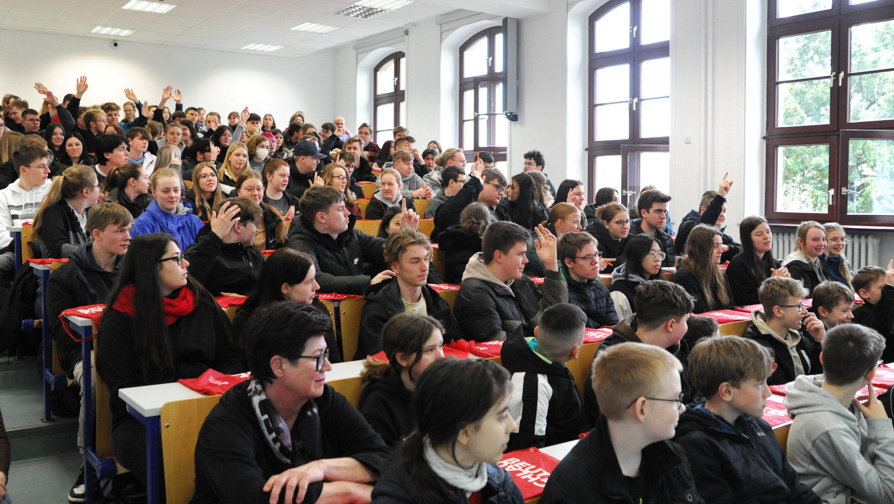 Großer Hörsaal mit vielen Schülern, die aufmerksam sitzen, einige heben die Hände, vermutlich zur Beantwortung von Fragen oder Teilnahme an einer Umfrage.