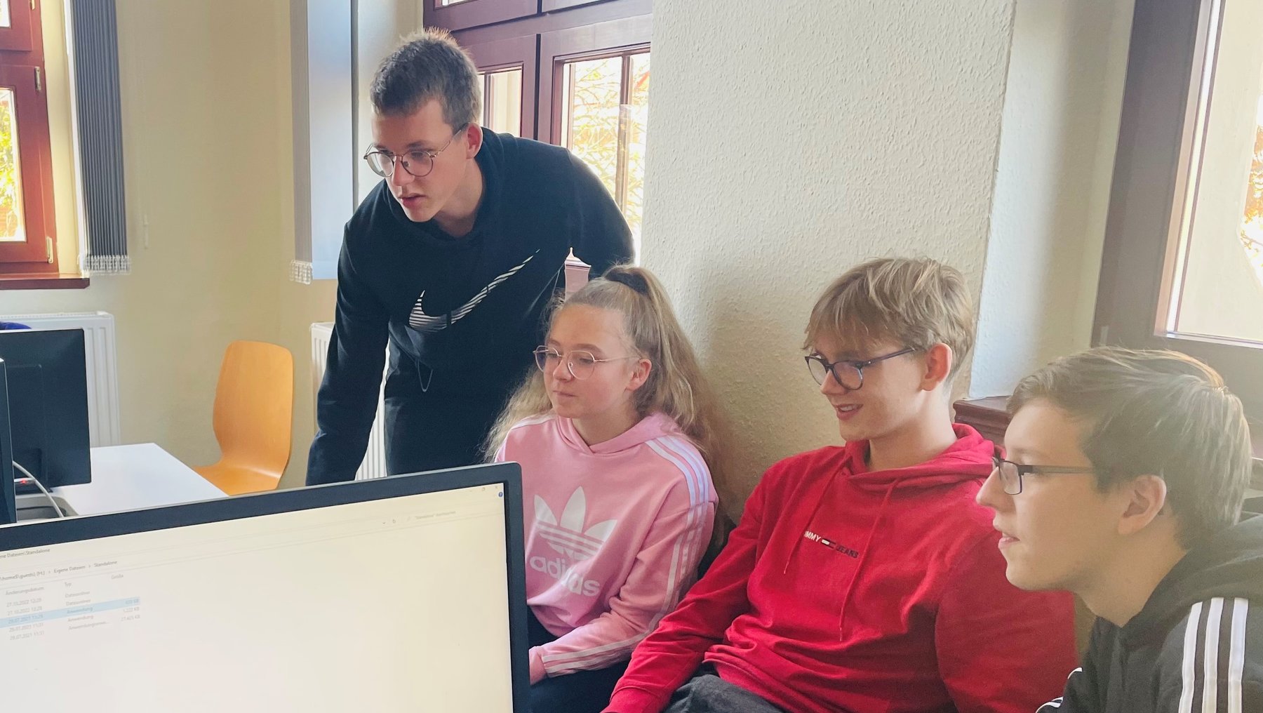 Ein männlicher Jugendlicher steht und erklärt drei Jugendlichen am Computer etwas, während diese konzentriert auf den Bildschirm schauen. Die Szene spielt in einem hellen Raum mit natürlichem Licht.
