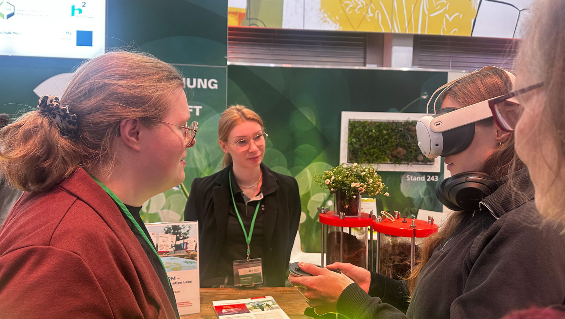 Drei Personen an einem Messestand: Eine Mitarbeiterin erklärt zwei Besucherinnen eine Virtual-Reality-Brille, die eine davon bereits trägt. Im Hintergrund sind Infomaterialien und ein Bildschirm mit Standnummer 243 zu sehen.