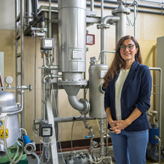 Eine Person mit Brille und blauem Blazer steht lächelnd vor einer technischen Anlage mit mehreren großen Metallbehältern, Rohrleitungen und Messgeräten in einer Industrieumgebung.