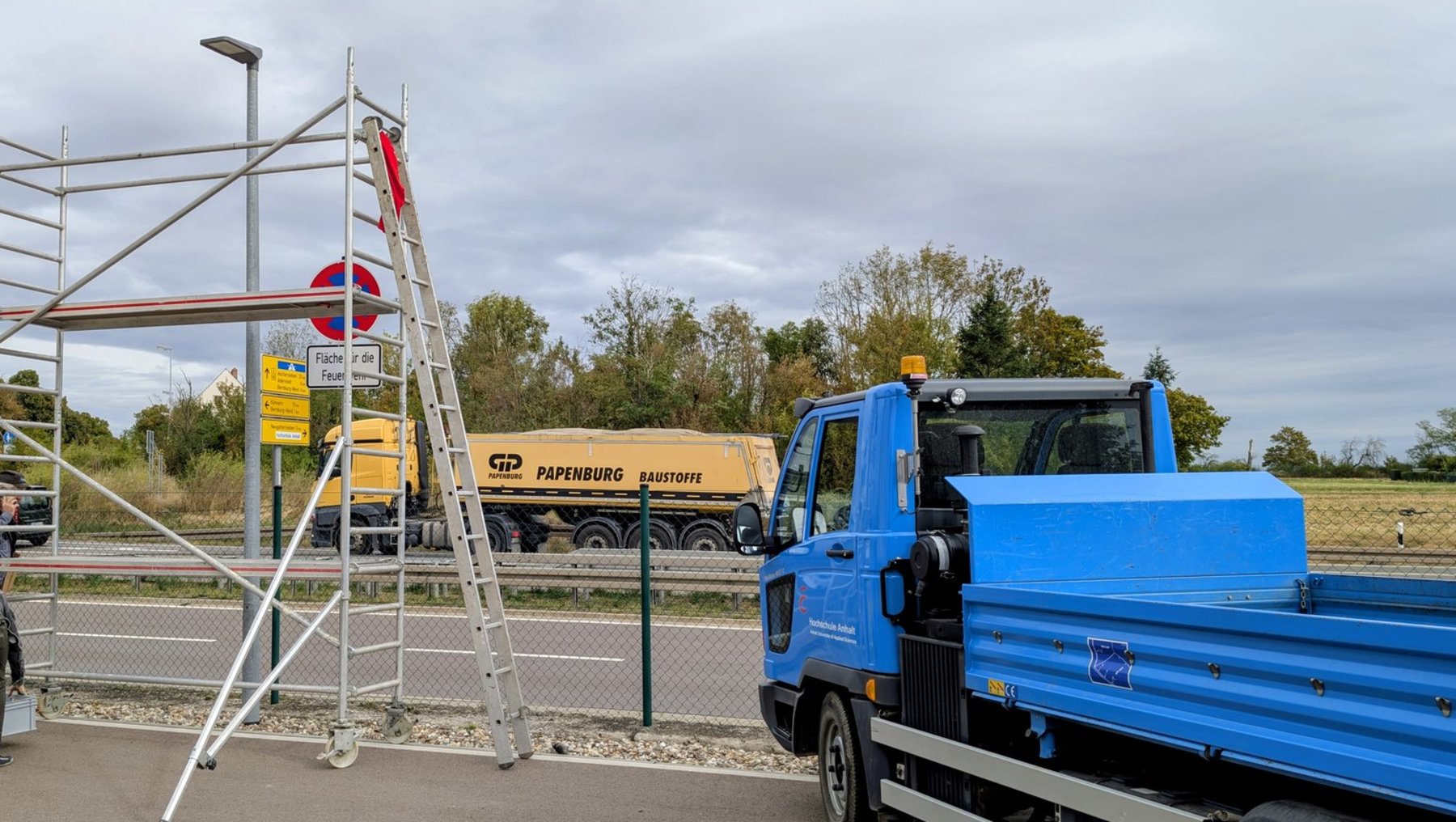 Blauer Lkw mit offener Ladefläche steht auf einer Straße. Im Hintergrund fährt ein gelber Müllwagen mit der Aufschrift „Abfallentsorgung“ vorbei. Eine Leiter lehnt an einem Zaun, daneben ein Schild mit Durchfahrtsverbot.