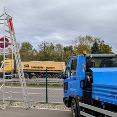 Blauer Lkw mit offener Ladefläche steht auf einer Straße. Im Hintergrund fährt ein gelber Müllwagen mit der Aufschrift „Abfallentsorgung“ vorbei. Eine Leiter lehnt an einem Zaun, daneben ein Schild mit Durchfahrtsverbot.