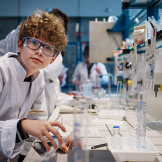 Ein junger Mensch in weißem Laborkittel und Schutzbrille führt konzentriert ein Experiment in einem Labor durch, umgeben von Reagenzgläsern, Flüssigkeiten und Laborgeräten.