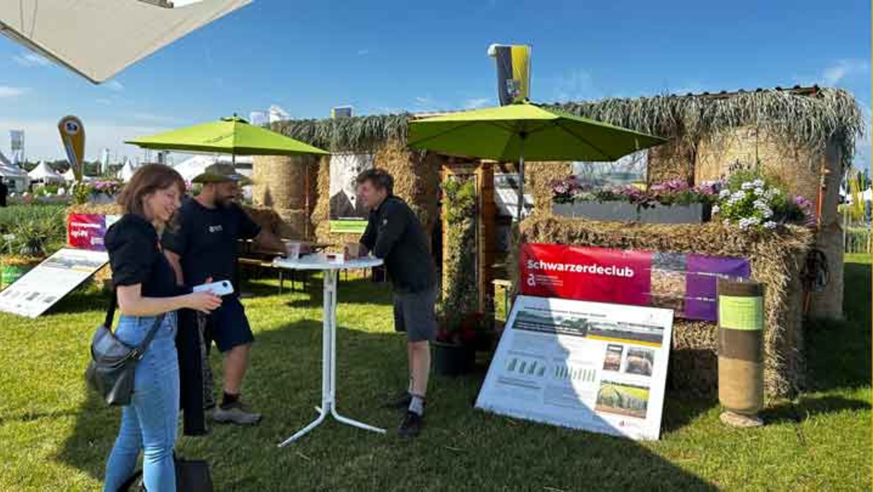 Drei Personen auf einer Messe oder einem Marktstand mit grüner Markise, der dem „Schwärzderclub“ zugeordnet ist. Sie stehen um einen Tisch mit Infomaterial und einer Infografik zu nachhaltigen Themen. Im Hintergrund sind Pflanzen und weitere Stände zu sehen.