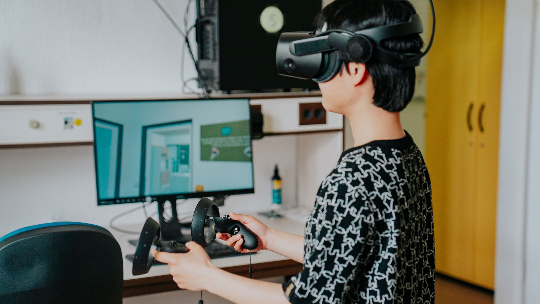 Eine Person nutzt eine Virtual-Reality-Brille und hält zwei VR-Controller in den Händen, während sie vor einem Bildschirm mit einer virtuellen Umgebung sitzt.