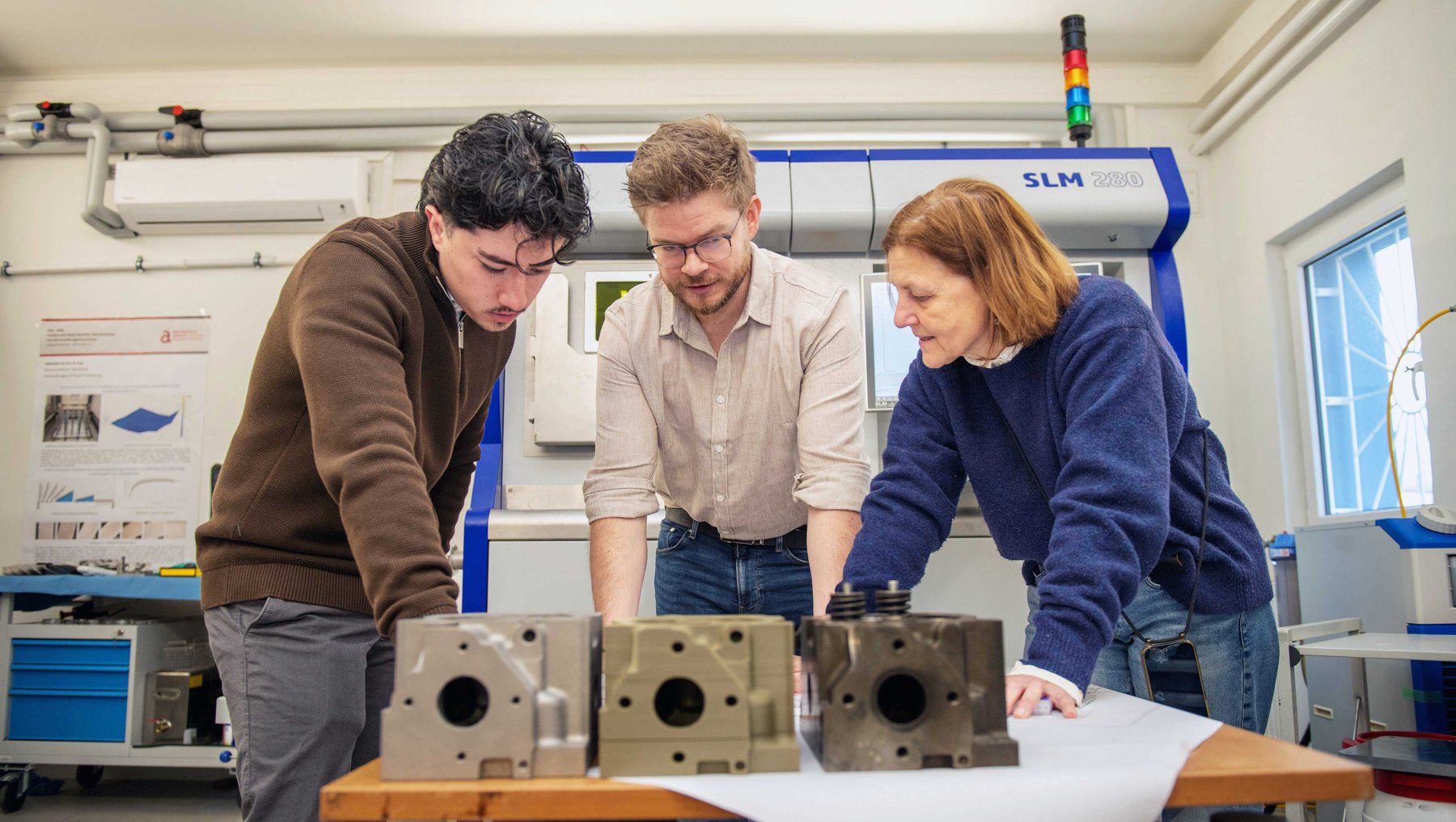 Drei Personen untersuchen gemeinsam drei metallene, komplex geformte Bauteile, die auf einem Tisch liegen, vor einem SLM-3D-Drucker in einer Werkstattumgebung in den Laboren der Hochschule Anhalt
