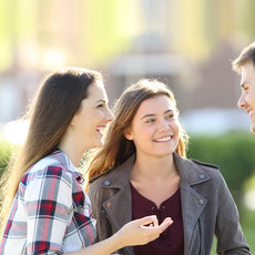 Three people are standing outside, smiling and engaged in conversation. The group consists of two women and one man. The women are wearing casual clothing, and the man is in a denim shirt. They appear to be enjoying a pleasant moment together in a sunny outdoor setting.