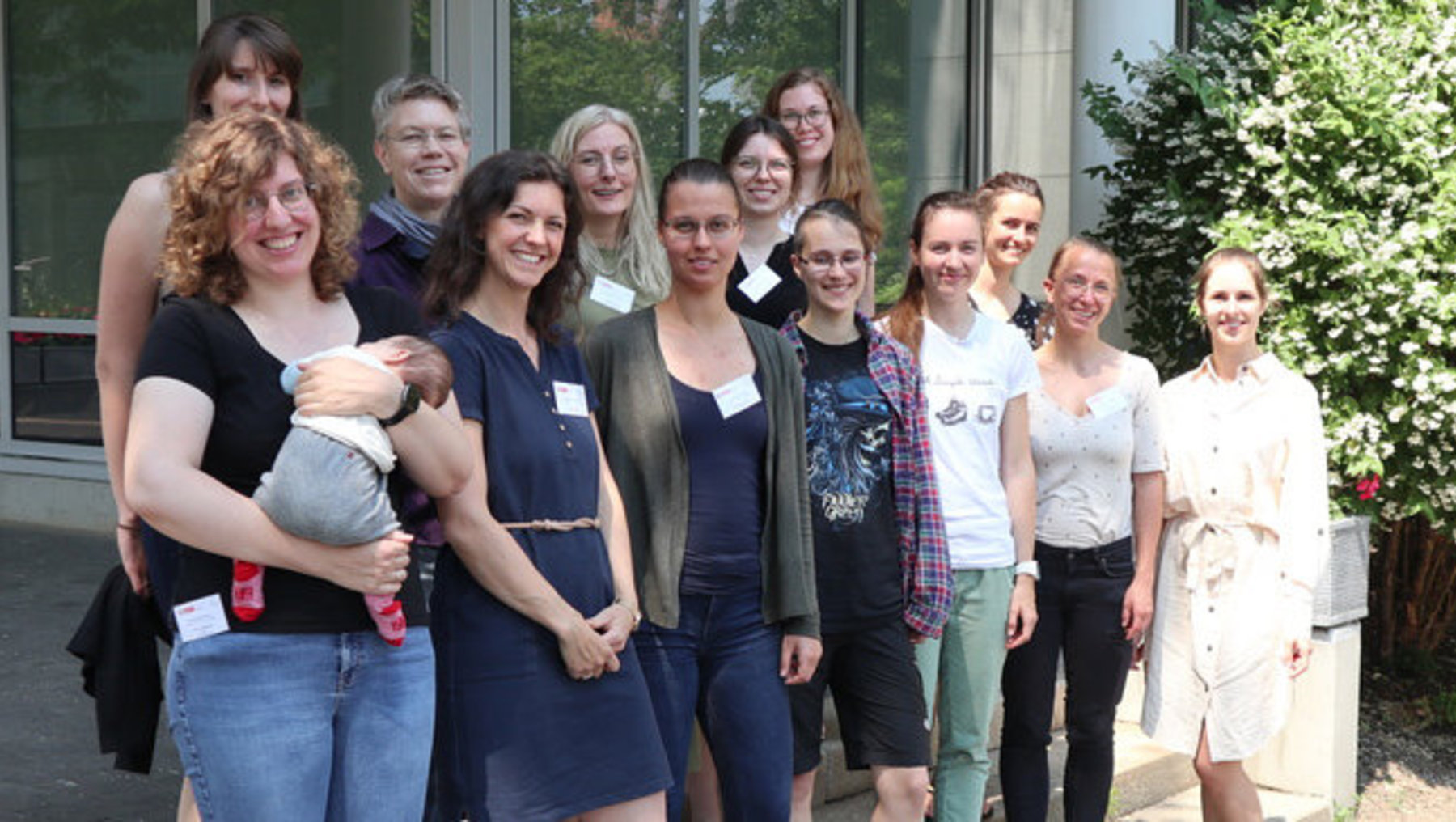 Eine Gruppe von Menschen steht vor einem Gebäude und lächelt in die Kamera. Die Gruppe besteht aus Erwachsenen, darunter eine Person, die ein Baby hält. Sie scheinen bei einem Gruppenfoto für einen besonderen Anlass zusammenzukommen.