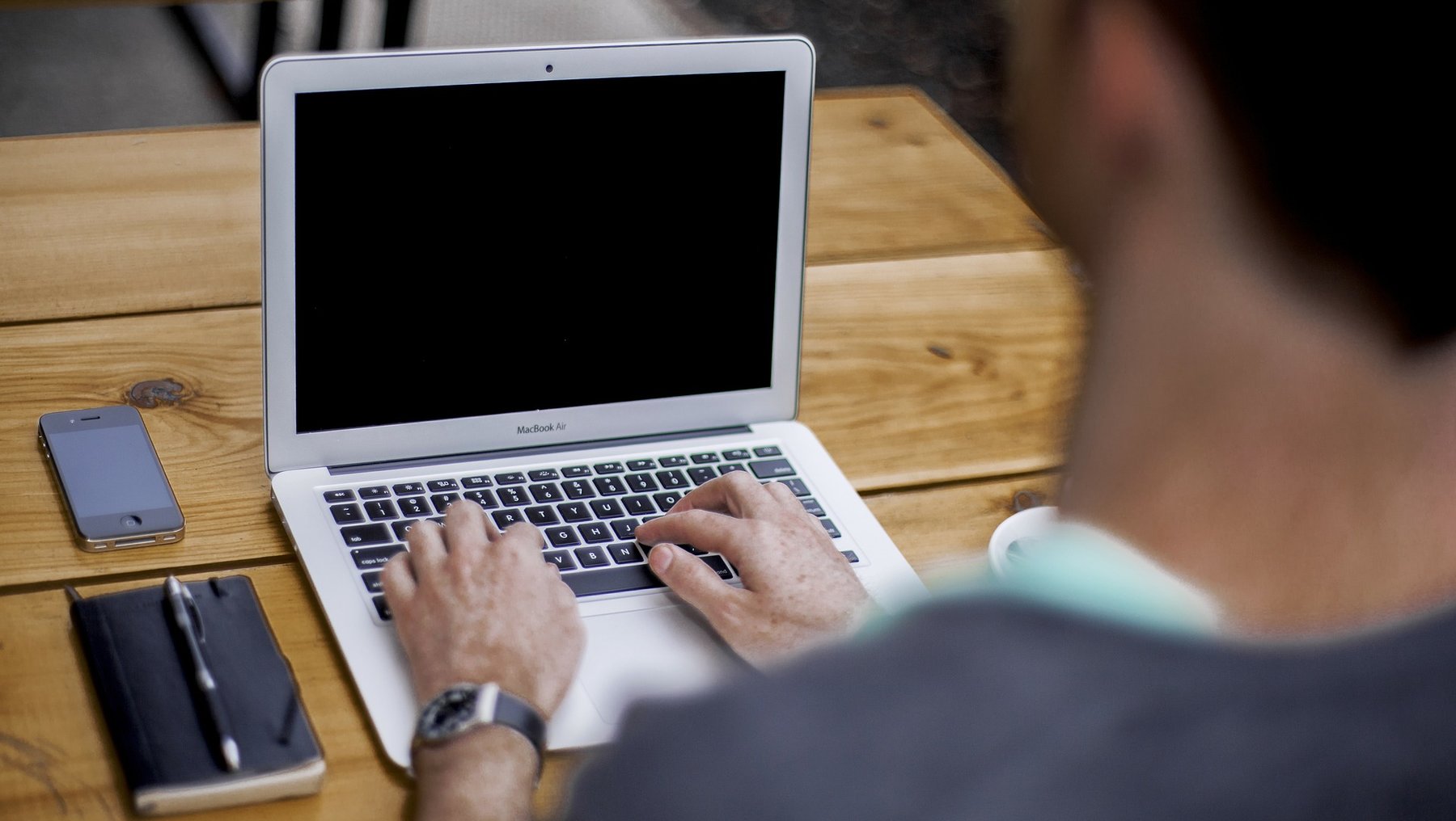 Ein Mensch arbeitet an einem Holztisch und tippt auf einem silbernen Laptop mit schwarze Tastatur und geschlossenem Display. Ein Smartphone liegt teilweise im Blickfeld auf der linken Seite des Tischs.