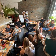 A group of people sitting in a cozy, well-lit room with modern decor. They are engaged in a discussion, some seated on couches and others on chairs. There are plants, a whiteboard, and a table with snacks and drinks, suggesting a casual meeting or team gathering.