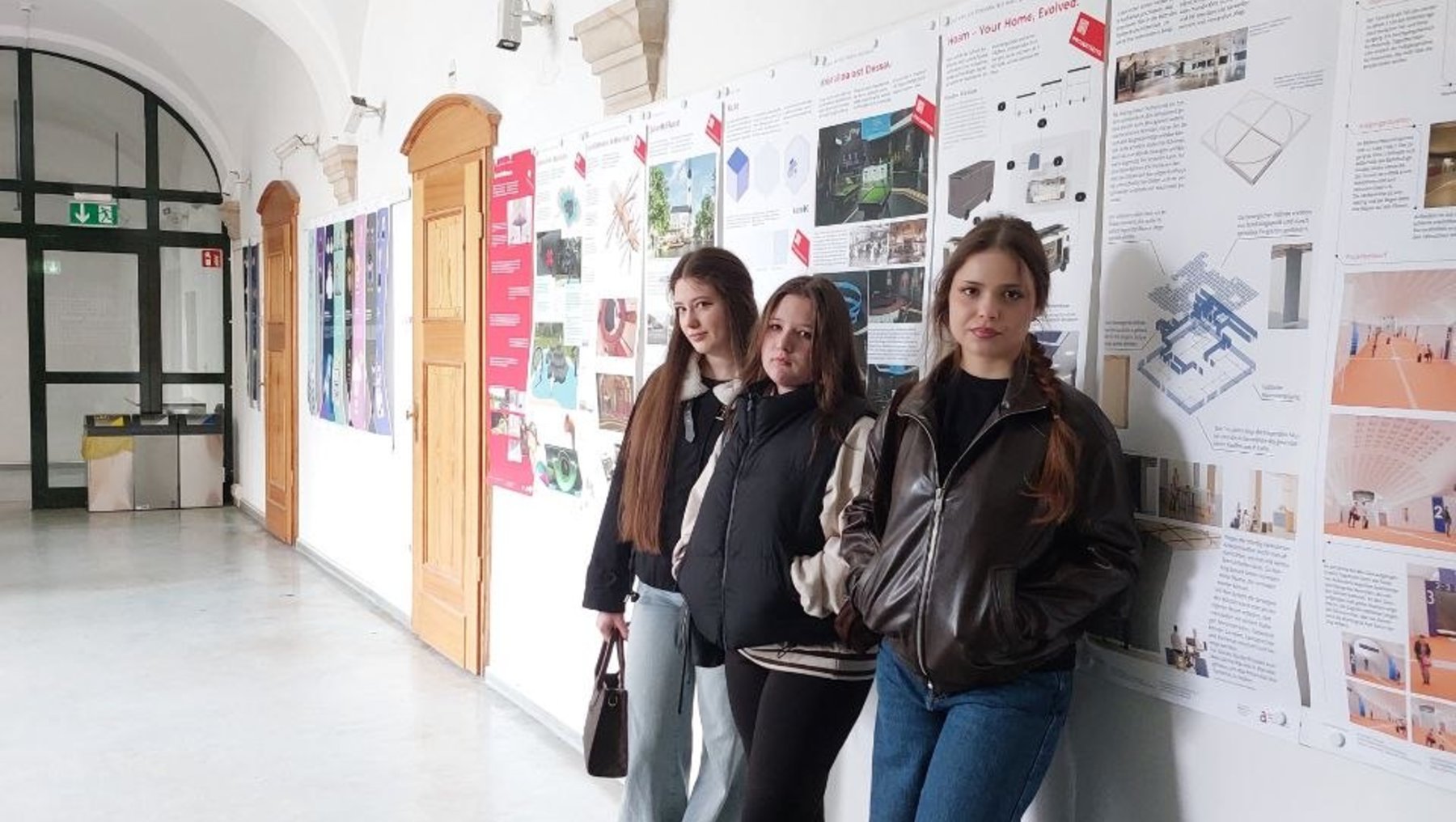 Drei Personen stehen in einem hellen Flur vor einer Wand mit mehreren Informationsplakaten, die Projekte oder Präsentationen zeigen. Die Plakate enthalten Texte, Grafiken und Fotos zu verschiedenen Themen.
