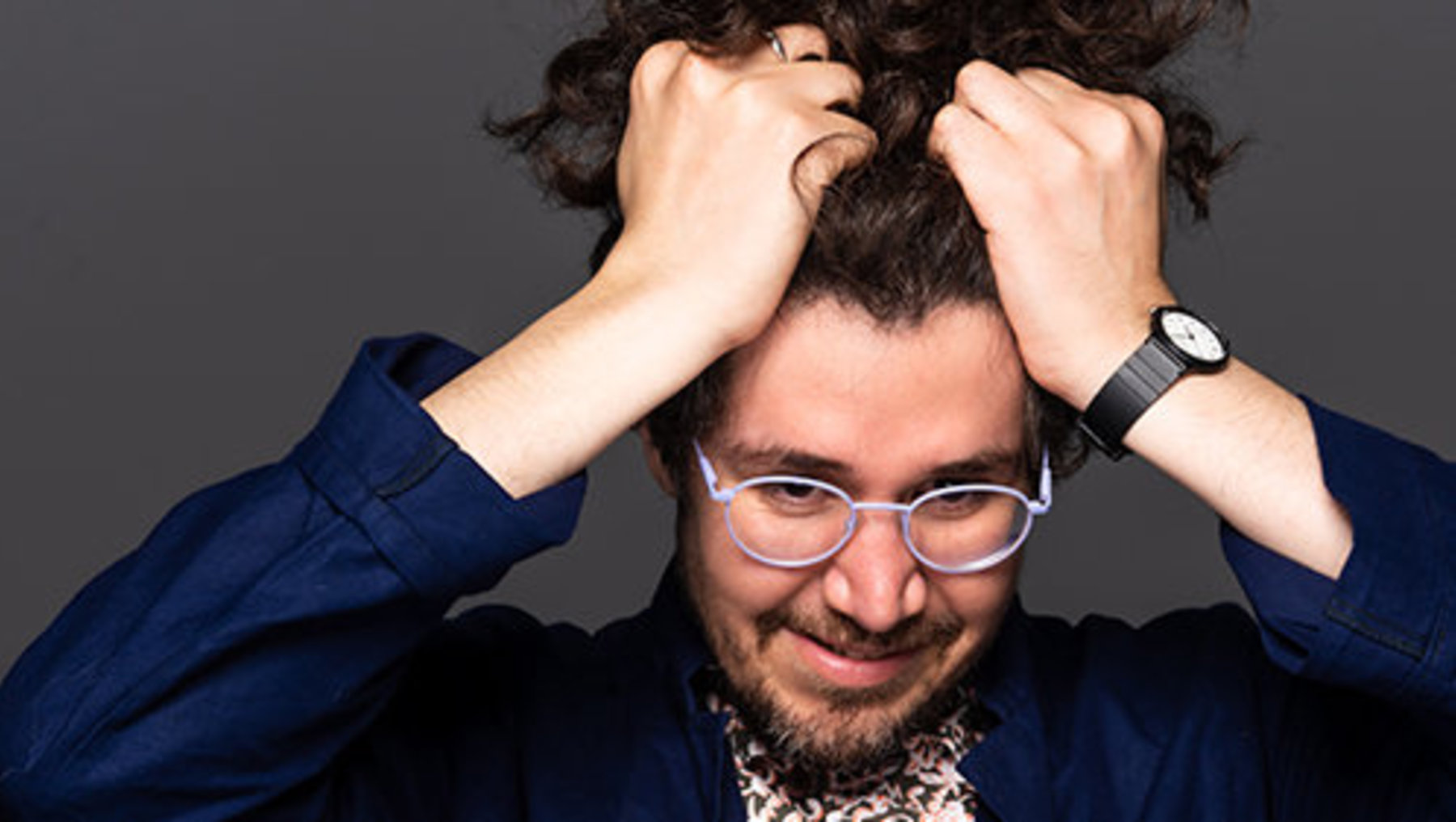 Ein Mann mit Brille und kurzem, lockigem Haar, der mit beiden Händen an den Kopf fasst, lächelnd und leicht verzweifelt wirkend. Er trägt eine blaue Jacke über einem gemusterten Hemd.