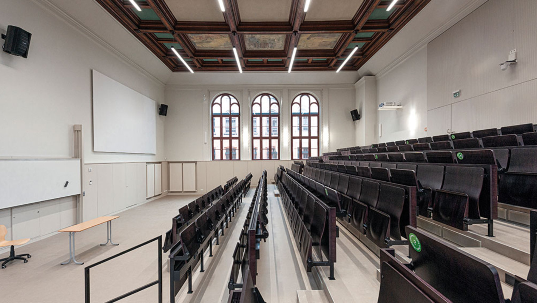 Ein leerer Hörsaal mit mehreren Reihen schwarzer Stühle, die zu einem Podium mit einem Whiteboard ausgerichtet sind. Große Bogenfenster lassen Licht herein, und die Decke hat eine Holzvertäfelung mit eingebauten Lichtern. Der Raum wirkt sauber und modern.