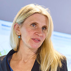 Eine Frau mit hellblonden, langen Haaren lächelt leicht zur Seite, trägt ein dunkles Oberteil und ein kleines Ohrschmuckstück. Sie wirkt freundlich und professionell.