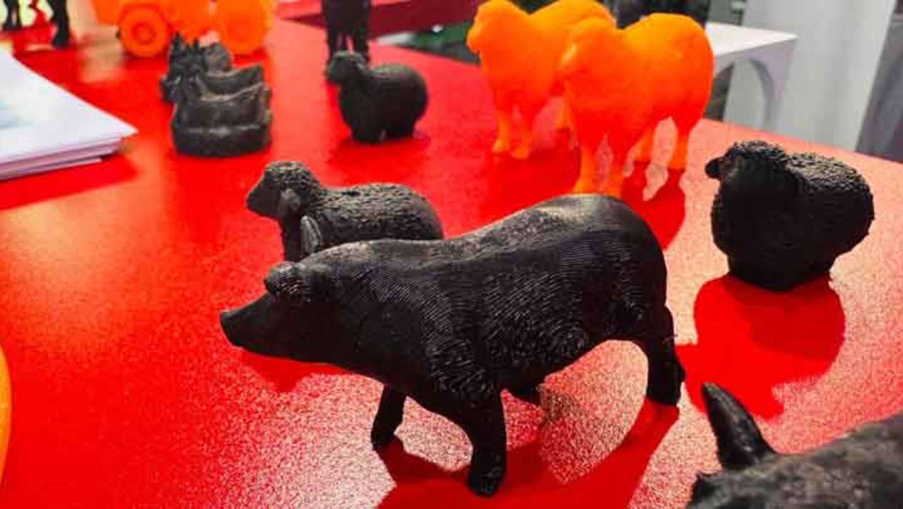 Drei kleine, schwarze Tierfiguren aus Ton oder Keramik auf einer roten Fläche: ein Schwein, ein Schaf und ein Hund, die aufrecht stehend oder sitzend angeordnet sind. Daneben weitere Tierfiguren in verschiedenen Farben und Materialien.