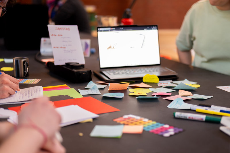 Ein Tisch mit bunten Karten und Post-its in verschiedenen Farben liegt verstreut. Eine Person hält einen Stift und notiert etwas auf einem Zettel. Ein Laptop zeigt eine Präsentation mit einem Diagramm. Kreatives Brainstorming oder Planung in einer Gruppe.
