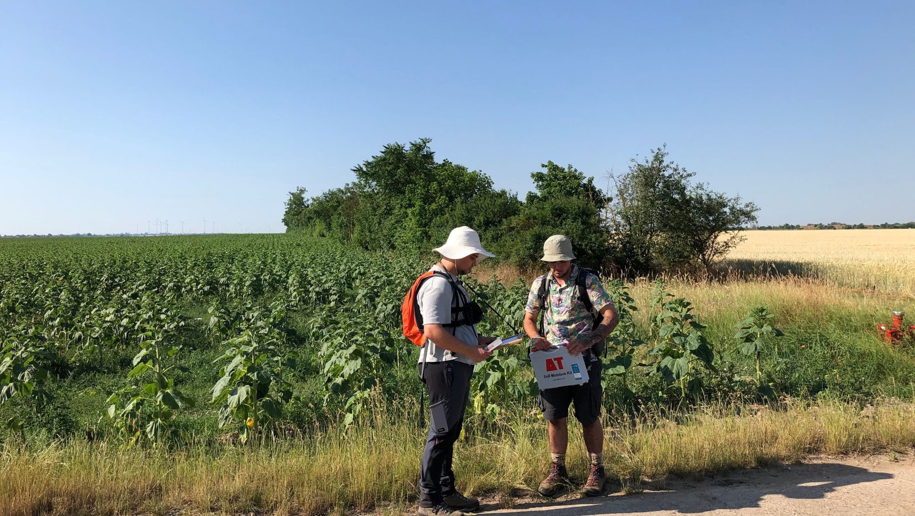 Zwei Personen in Schutzbekleidung und Hüten stehen auf einem Feldweg. Sie besprechen sich und halten Messwerkzeuge in der Hand. Im Hintergrund ist ein Feld mit gelbem Raps zu sehen.