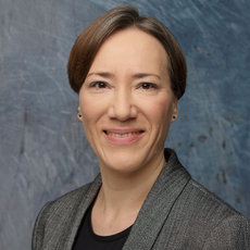 A woman with short brown hair, wearing a black top and a gray blazer, smiles at the camera. She has small earrings and is set against a blue textured background. The image conveys a professional and approachable demeanor.
