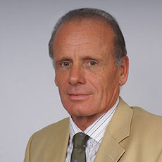 A man with short gray hair wearing a beige blazer, a striped dress shirt, and a green tie. He is looking directly at the camera with a neutral expression. The background is plain and light-colored.