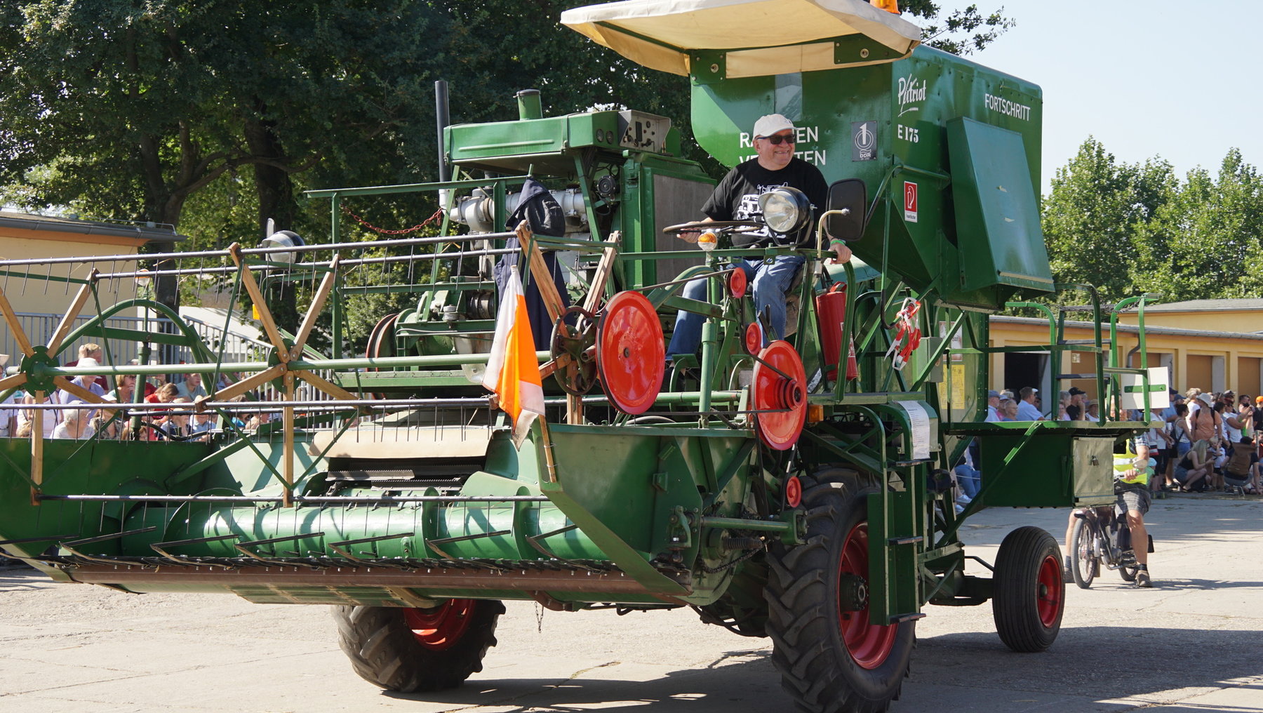 Grüner historischer Mähdrescher der Marke Pfaff mit rot-schwarzen Reifen, der von einer Person gesteuert wird. Das Fahrzeug ist mit einem großen Schneidwerk und Förderband ausgestattet und steht auf einer öffentlichen Fläche mit Zuschauern im Hintergrund.