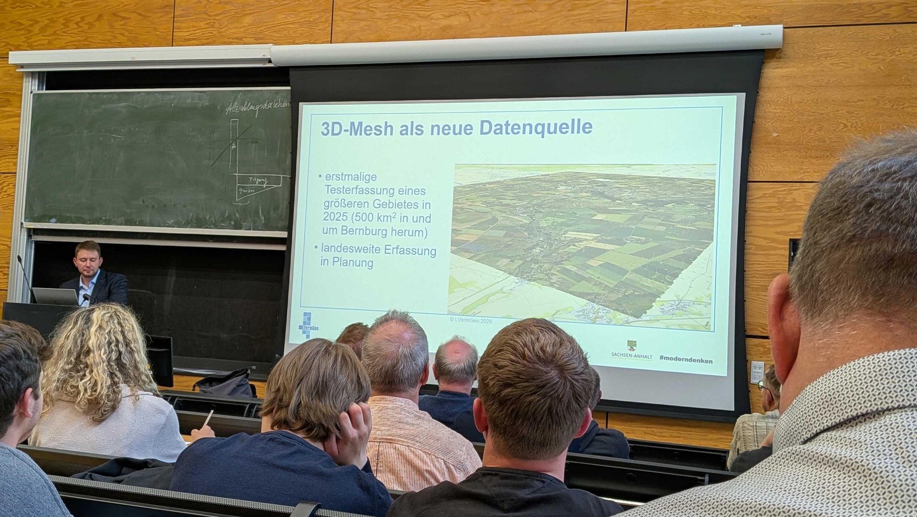 Vortragsraum mit Publikum, das einem Referenten zuhört. Auf der Leinwand wird eine Präsentation mit dem Titel „3D-Mesh als neue Datenquelle“ gezeigt, darunter ein 3D-Modell eines Geländes mit Gebäuden und Vegetation.