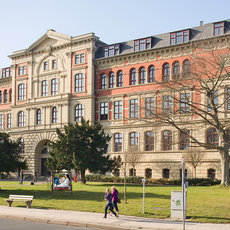 A large, historic building with a red and beige facade, featuring numerous windows and a central archway entrance. The building is surrounded by a well-maintained lawn with a few trees and benches. Two people are walking on a path in front of the building.