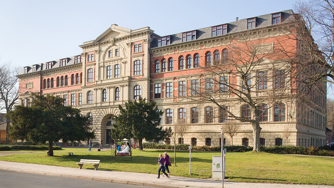 Ein großes, historisches Gebäude mit roten und hellen Steinfassaden, vielen Fenstern und einem zentralen Eingang. Vor dem Gebäude befindet sich ein gepflegter Rasen mit Bäumen, Bänken und zwei Personen, die spazieren gehen. Die Szene wirkt friedlich und ist von einem klaren Himmel umgeben.
