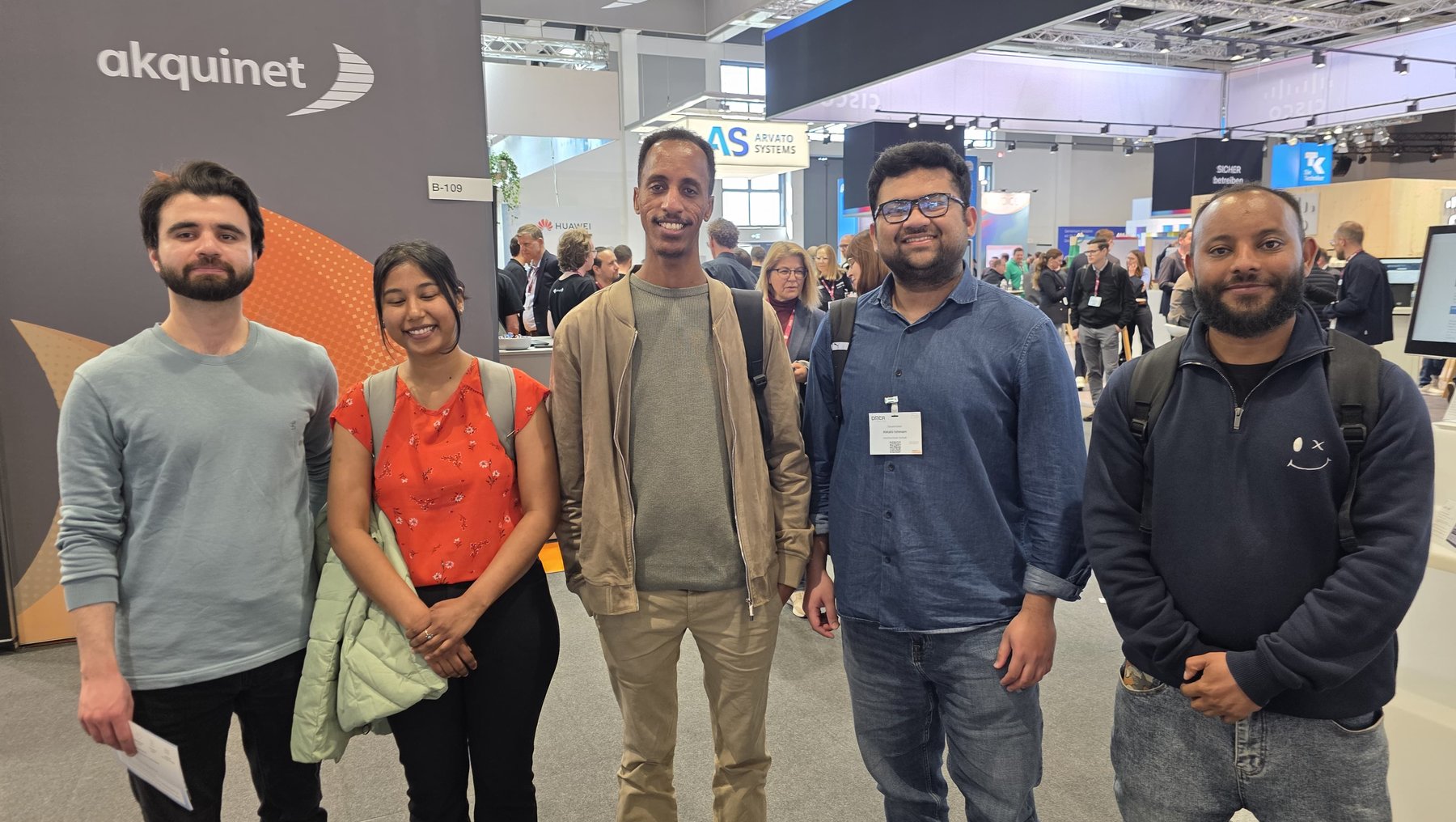 Fünf Personen stehen lächelnd auf einer Messe vor einem Stand mit dem Logo „akquinet“ und im Hintergrund weiteren Ausstellern wie „Cisco“. Sie tragen verschiedene Kleidungsstücke und wirken entspannt.