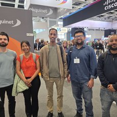 Fünf Personen stehen lächelnd auf einer Messe vor einem Stand mit dem Logo „akquinet“ und im Hintergrund weiteren Ausstellern wie „Cisco“. Sie tragen verschiedene Kleidungsstücke und wirken entspannt.