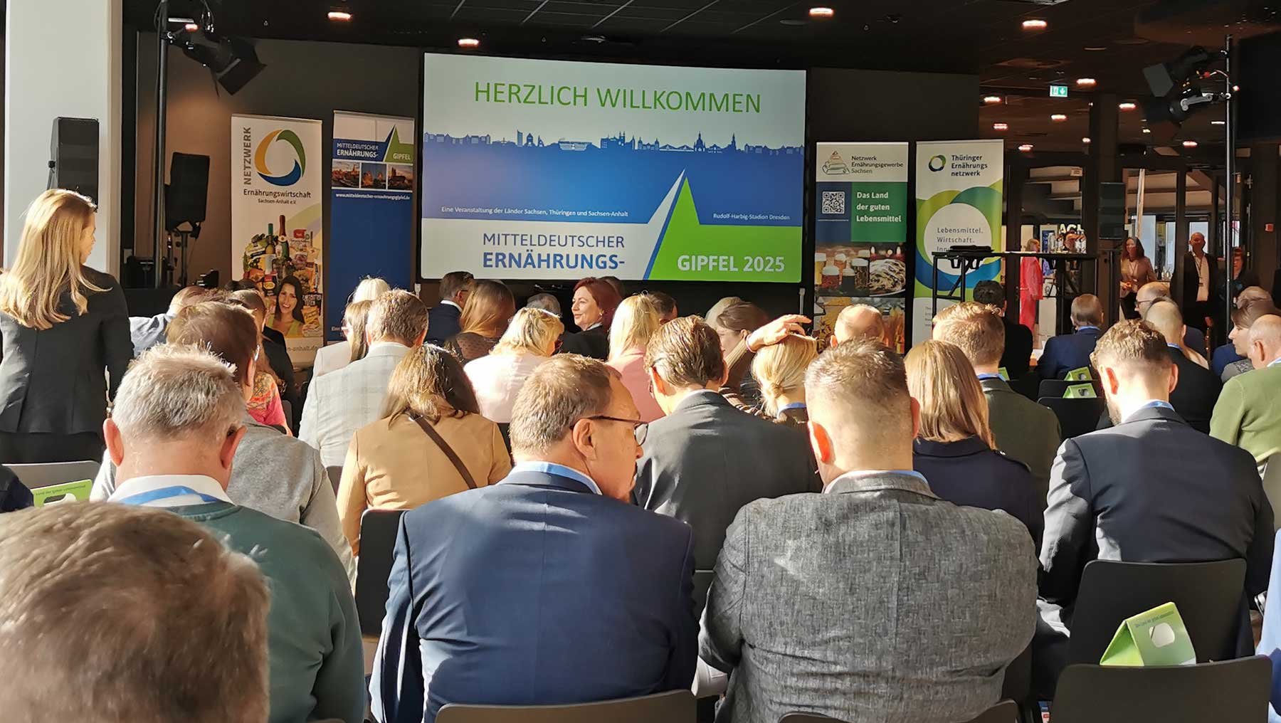 Großer Saal mit vielen sitzenden und stehenden Personen bei einer Veranstaltung. Im Hintergrund ein Banner mit der Begrüßung „Herzlich willkommen“ und dem Titel „Mitteldeutscher Ernährungsgipfel 2025“