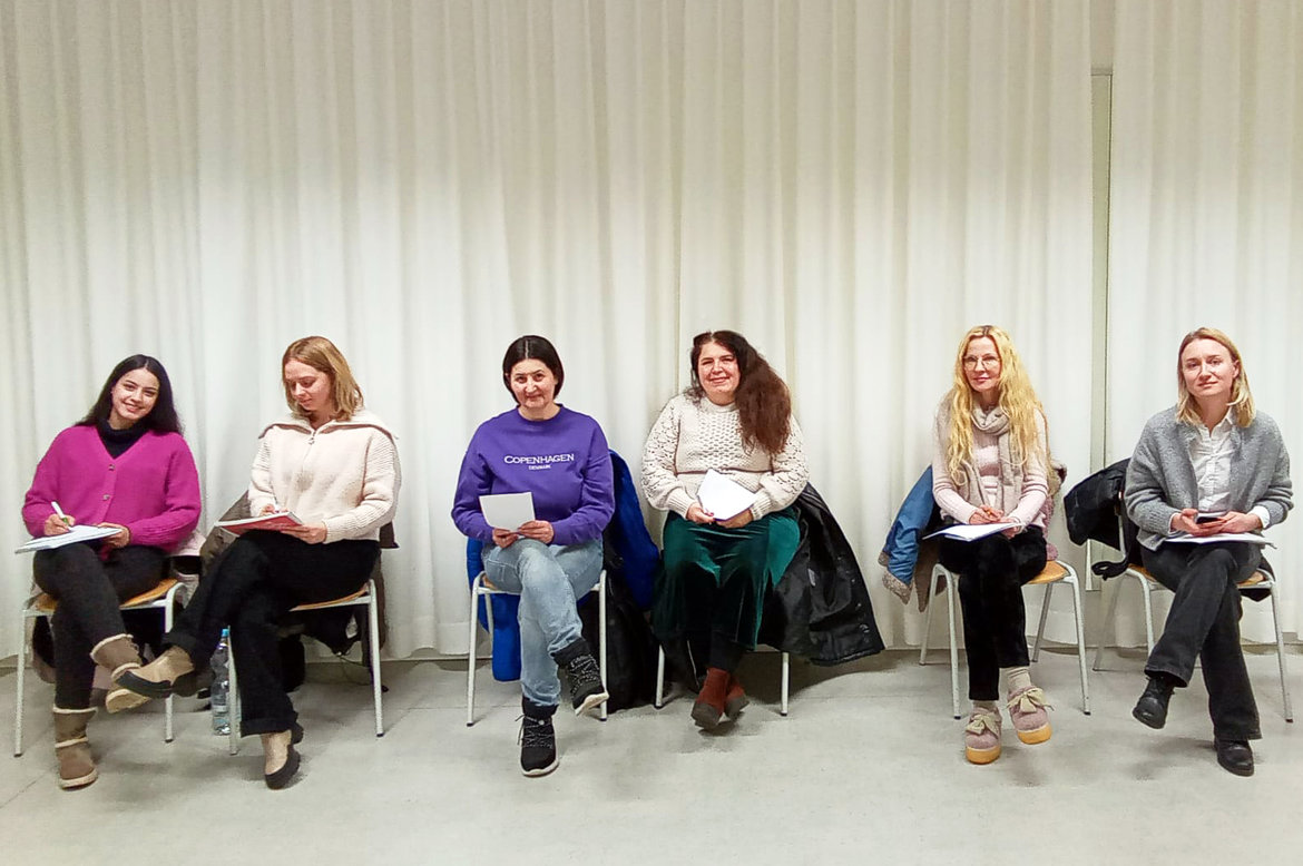 Fünf Menschen, wahrscheinlich Teilnehmerinnen einer Diskussion oder Arbeitsgruppe, sitzen mit Notizblöcken auf ihren Schoßen auf Stühlen in einem Innenraum – drei mit langem, zwei mit kurzem Haar – und wirken konzentriert.