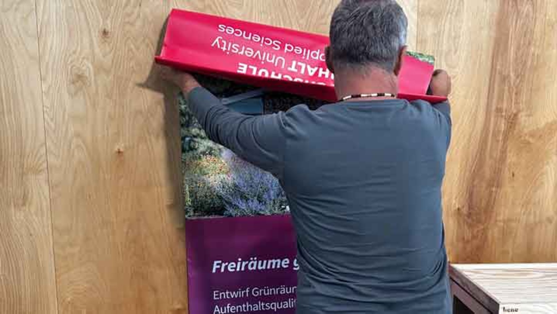 Ein Mann befestigt ein großes Plakat an einer Holzvertäfelung. Das Plakat zeigt eine blühende Pflanze und den Text Freiräume gestalten mit Bezug auf Entwurfsprozesse für Grünräume und Aufenthaltsqualität an der HAW Hamburg, Fakultät Life Sciences.