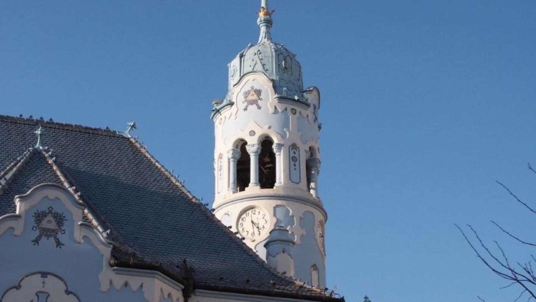 Barocke Kirche mit weißer Fassade und blauen Verzierungen, Turm mit Uhr und Kreuz auf dem Dach, klarer Himmel im Hintergrund.
