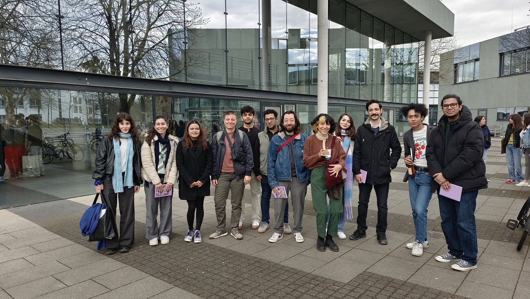 Gruppe von 14 Personen, die vor einem modernen Gebäude stehen, lächeln und für ein Gruppenfoto posieren. Alle tragen Winterkleidung und halten teilweise Flyer oder Plakate.