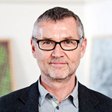 A middle-aged man with short gray hair and glasses, wearing a dark blazer and a light-colored shirt, looking directly at the camera with a neutral expression. The background is blurred, indicating an indoor setting.