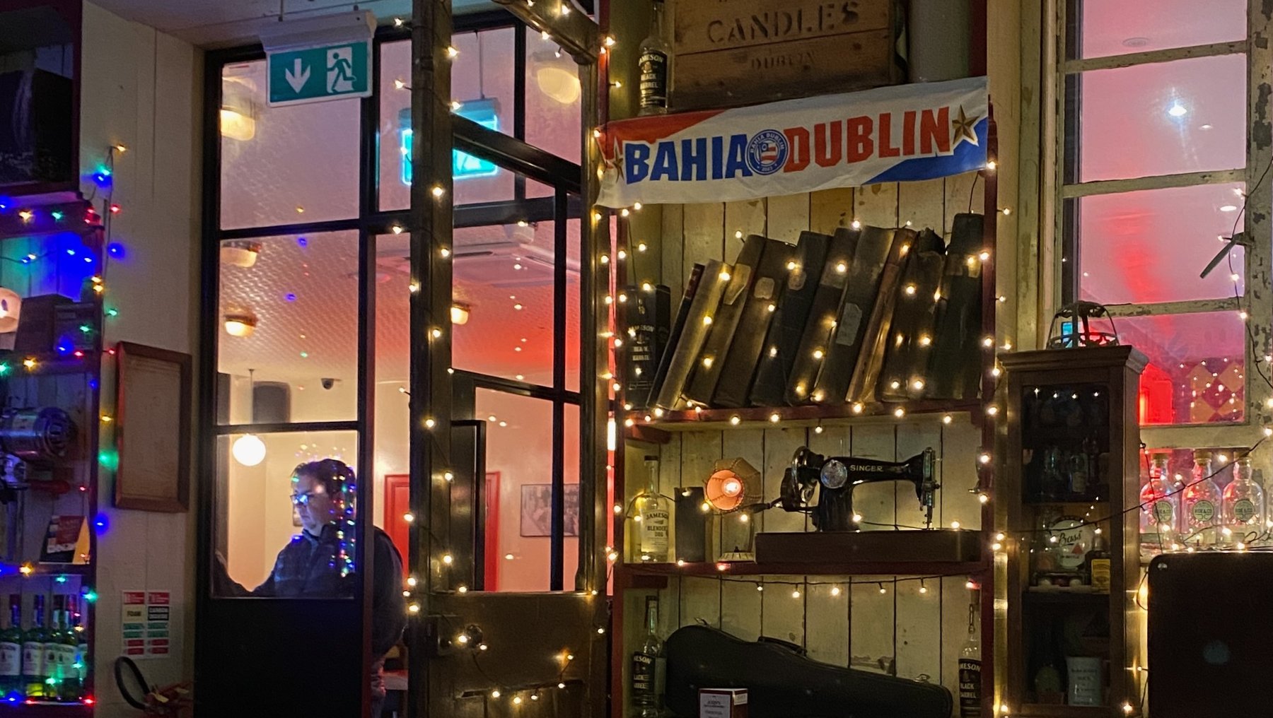 Gemütliche Bar mit dem Namen Bahia Dublin, geschmückt mit warmem Lichterketten-Dekor. Im Inneren sind Theke mit Flaschen, ein Tresen mit Stühlen und eine Person hinter der Bar zu erkennen.