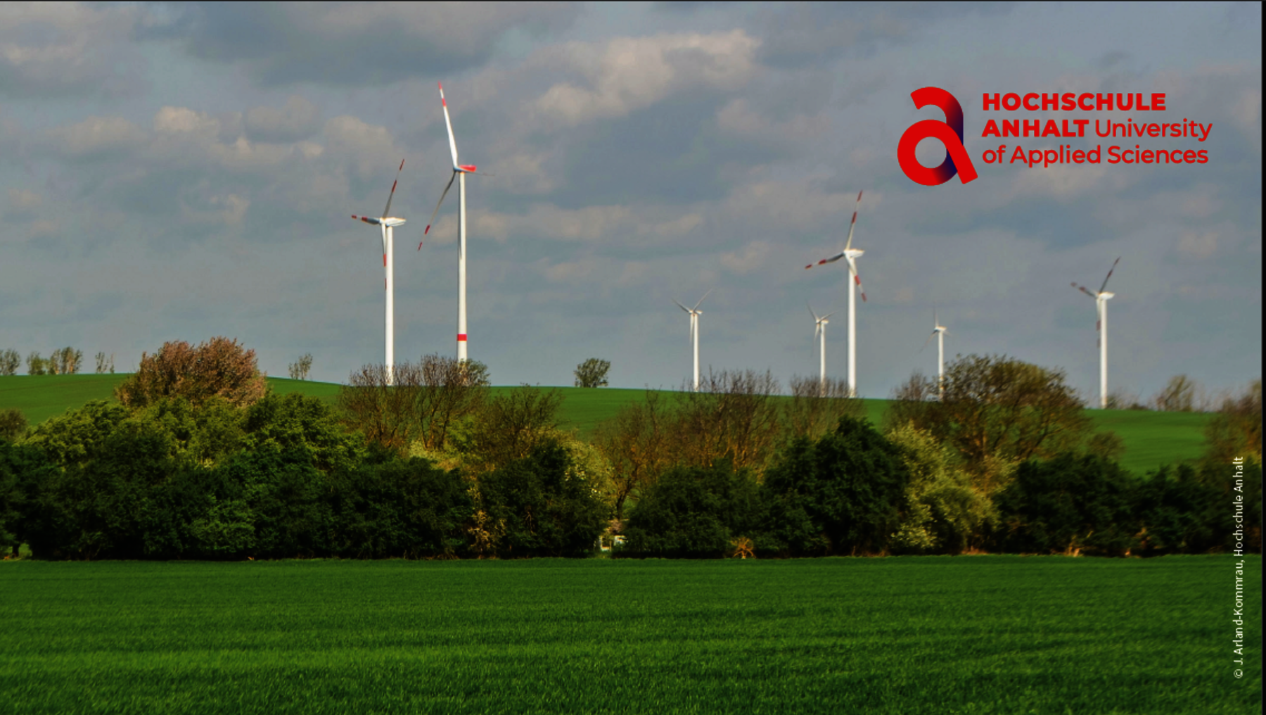 Grüne Landschaft mit sanften Hügeln, auf denen mehrere Windkraftanlagen in einer Reihe stehen. Im Hintergrund bewölkter Himmel mit Sonnenlicht.