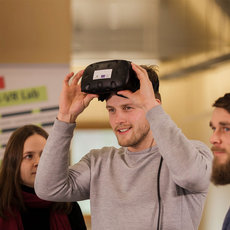 A man is wearing a virtual reality headset while two other people observe him. They appear to be in a lab or classroom setting, possibly demonstrating or experiencing virtual reality technology.