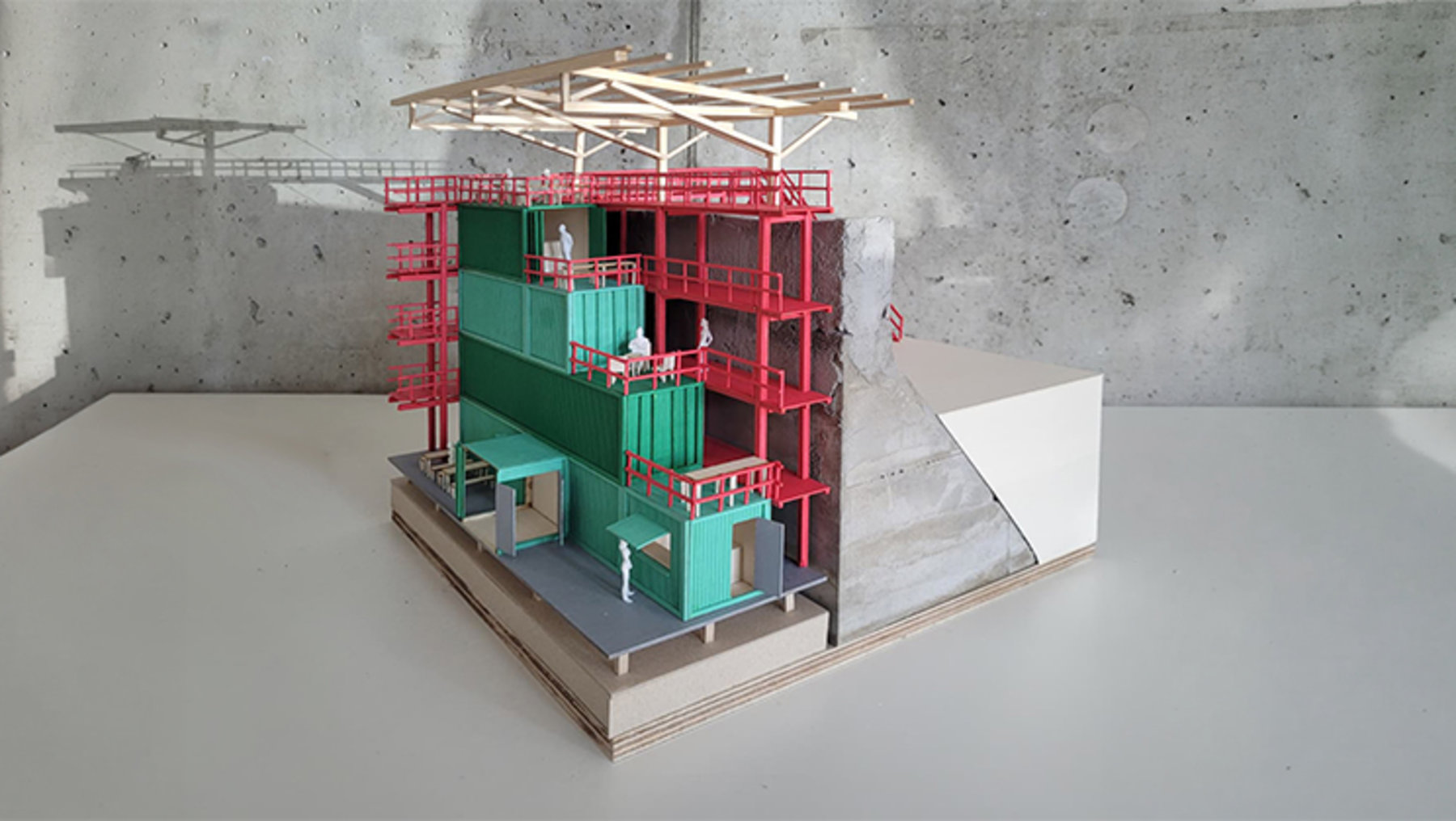 Modell eines mehrstöckigen Gebäudes im Rohbau mit roter Gerüstkonstruktion, grünen Außenwänden und sichtbaren Betonfundamenten. Im Hintergrund ein Teil einer Betonwand und eine kleine Brücke oder Plattform.