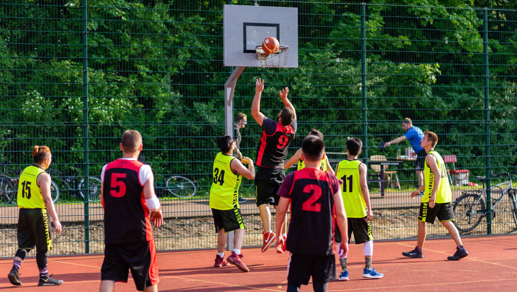 Ein Basketballspiel im Freien. Ein Spieler in rotem Trikot mit der Nummer 9 springt und wirft den Ball zum Korb, während andere Spieler in gelben und roten Trikots versuchen, den Wurf zu blockieren oder zu verteidigen.