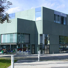 "Modernes Gebäude mit grauer Fassade und großen Glasfronten. Vor dem Gebäude befindet sich ein Gehweg mit einigen Personen. Im Hintergrund sind Bäume und ein klarer Himmel zu sehen."
