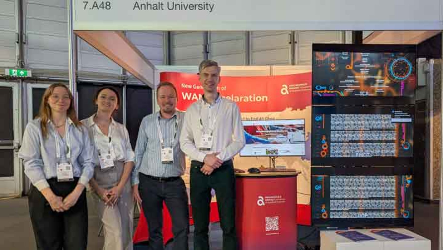 Vier Personen stehen lächelnd vor einem Messestand der Anhalt University mit dem Thema Wasserstoffforschung. Der Stand zeigt eine Präsentation zu Wasserstoff-Energie und -Anwendungen, darunter ein Poster mit technischen Darstellungen und einem Logo des Projekts.
