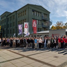 Große Gruppe von Studierenden versammelt sich vor einem grünen Hochschulgebäude der Hochschule Anhalt. Einige halten Fahnen mit der Zahl 6 und dem Logo der Hochschule. Sonniges Wetter, Bäume mit gelbem Laub im Hintergrund.