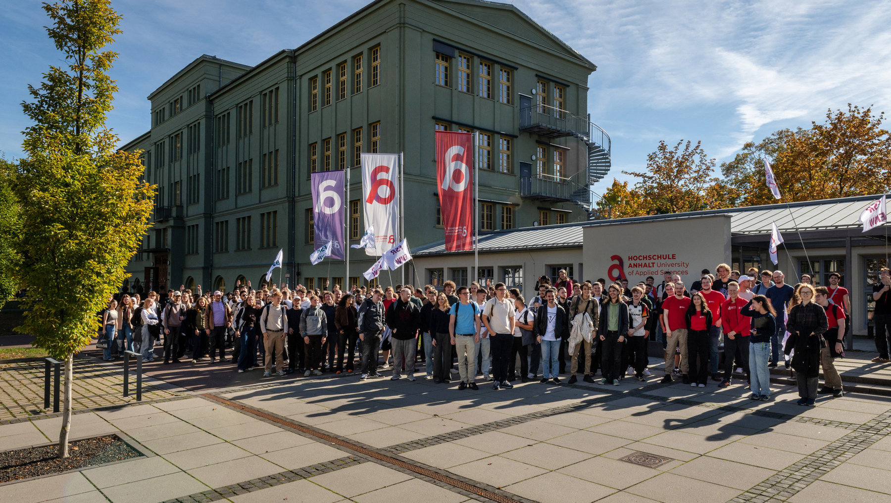 Große Gruppe von Studierenden versammelt sich vor einem grünen Hochschulgebäude der Hochschule Anhalt. Einige halten Fahnen mit der Zahl 6 und dem Logo der Hochschule. Sonniges Wetter, Bäume mit gelbem Laub im Hintergrund.