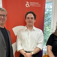 Drei Personen stehen lächelnd nebeneinander vor einem roten Banner mit dem Logo der Hochschule Aalen. Der Mann links trägt eine graue Jacke und Jeans, der Mann in der Mitte ein weißes Hemd, die Frau rechts ein dunkelblaues Shirt mit Logo und schwarze Hose.